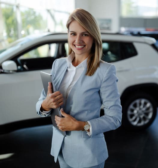 Female salesperson at a car showroom, holding a laptop and smiling to the camera.